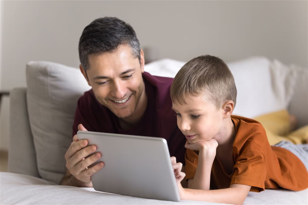 Vater mit Sohn liegen auf der Couch und schauen in einen Laptop.