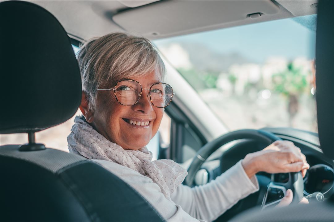 &Auml;ltere Frau sitzt l&auml;chelnd am Steuer eines Autos