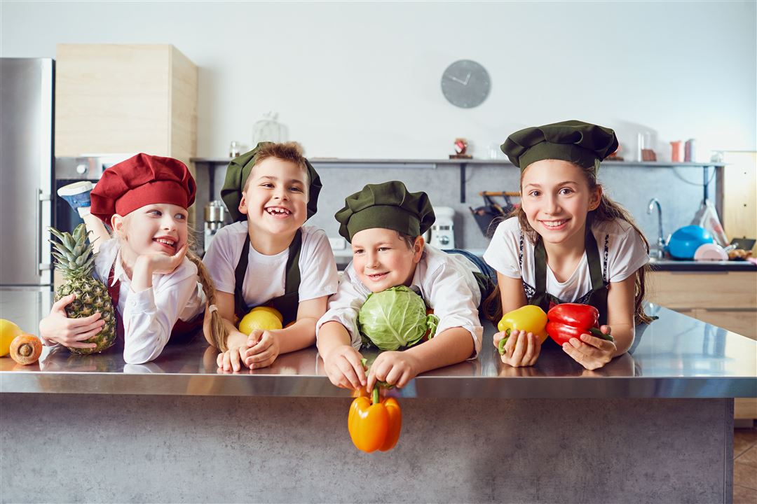 3 Kinder mit Kochm&uuml;tzen und Gem&uuml;se in den H&auml;nden, lachend auf K&uuml;chenarbeitsplatte.
