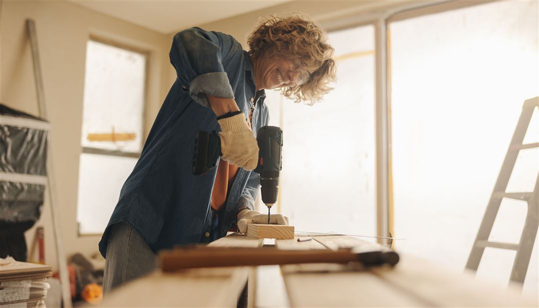 Ein Zimmer wird renoviert. Eine Frau mit Schutzbrille und Arbeitshandschuhen bohrt mit einer Bohrmaschine ein Loch in ein St&uuml;ck Holz.